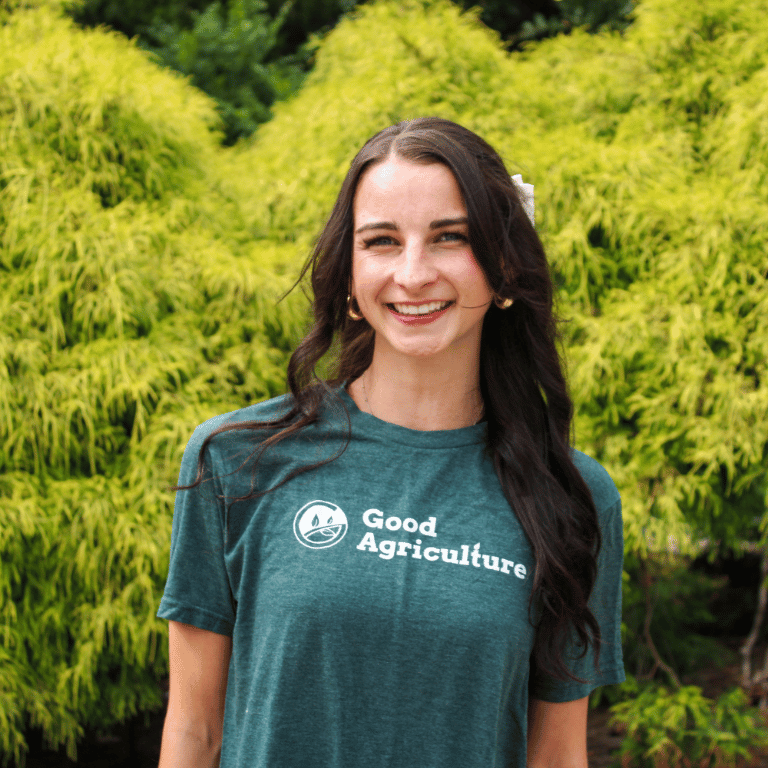 Lily from the Good Agriculture marketing team wearing a Good Agriculture shirt outdoors, smiling in front of greenery during a farm and agriculture brand photoshoot.
