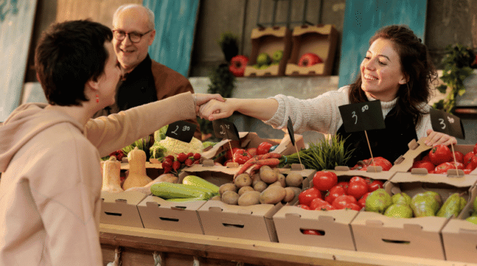 Farmers market vendor greeting a customer while selling fresh local produce, including tomatoes, potatoes, strawberries, and squash.