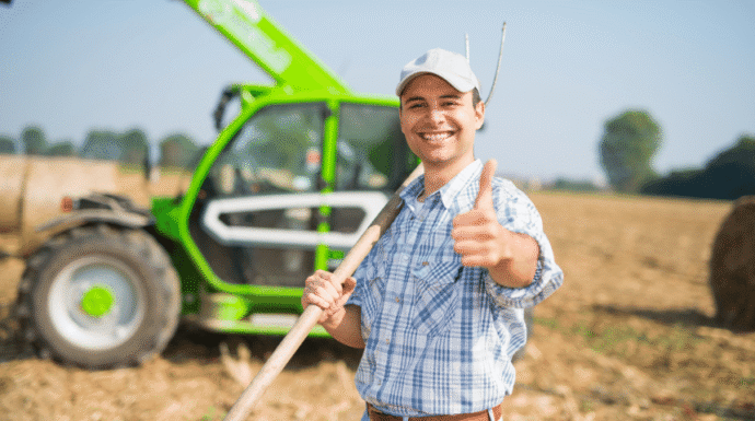 A farmer smiling and giving a thumbs-up while standing in a field beside a tractor, representing farming and agricultural work.