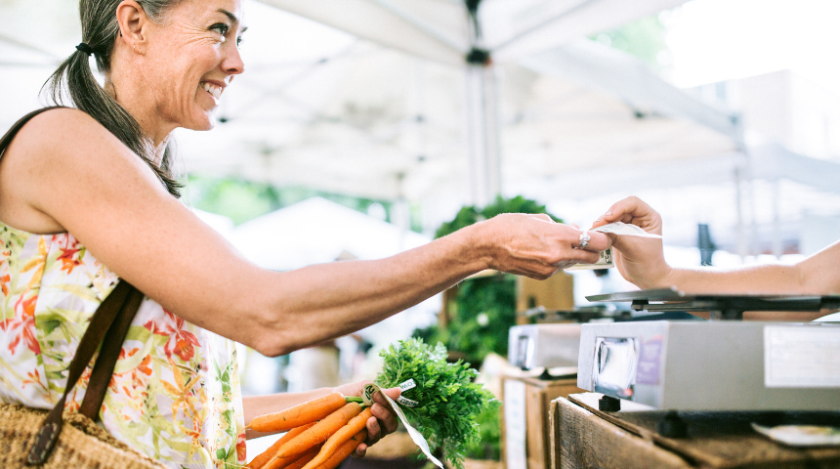 A customer purchases fresh carrots at a farmers market, handing cash to a vendor while holding a bunch of carrots.